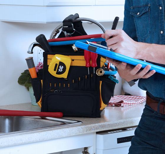 person holding a clipboard and work tools standing near an organized tool bag in a kitchen setting featuring five essential plumbing tools